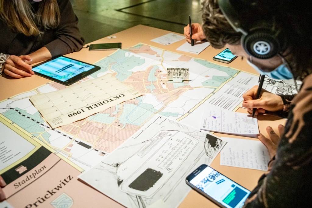 Der Inhalt eines Tischs ist zu sehen: Smartphones, ein Stadtplan der Stadt Prückwitz, bedruckte Blätter und die Hände von drei Personen, die um den Tisch herumsitzen. Zwei der Personen machen sich Notizen.