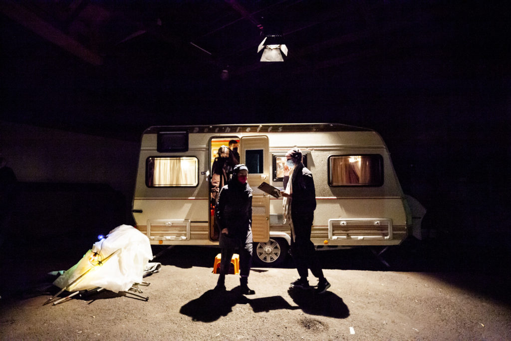 Ein von einem Scheinwerfer beleuchteter Wohnwagen, aus dem vier Menschen mit FFP2-Masken heraustreten.