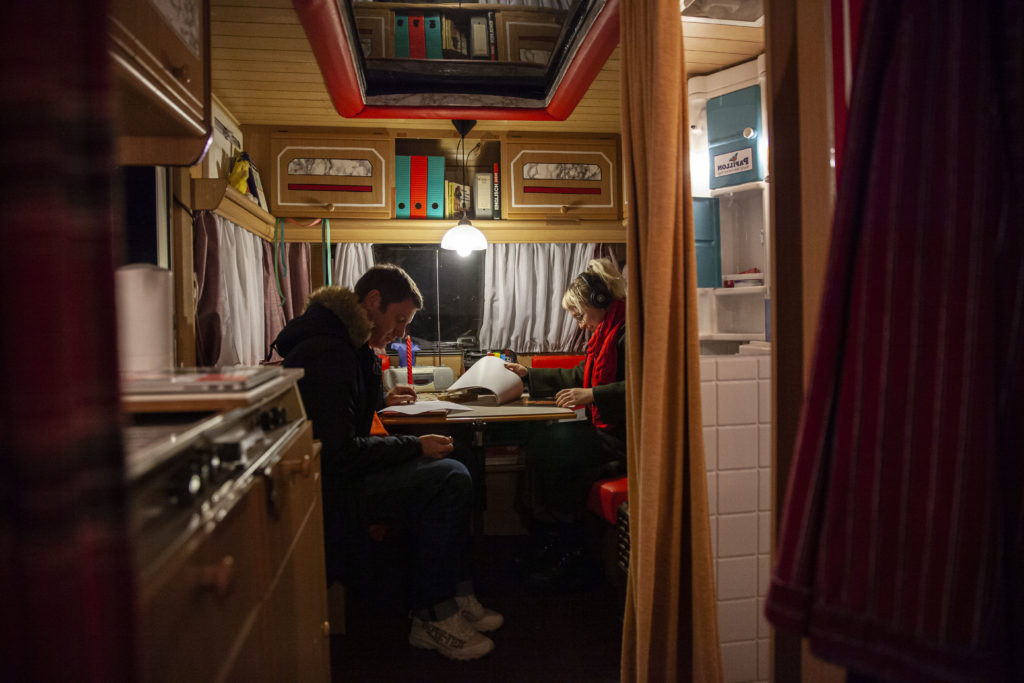 Zwei Menschen sitzen in einem Wohnwagen an einem Tisch und lesen bedruckte Blätter.