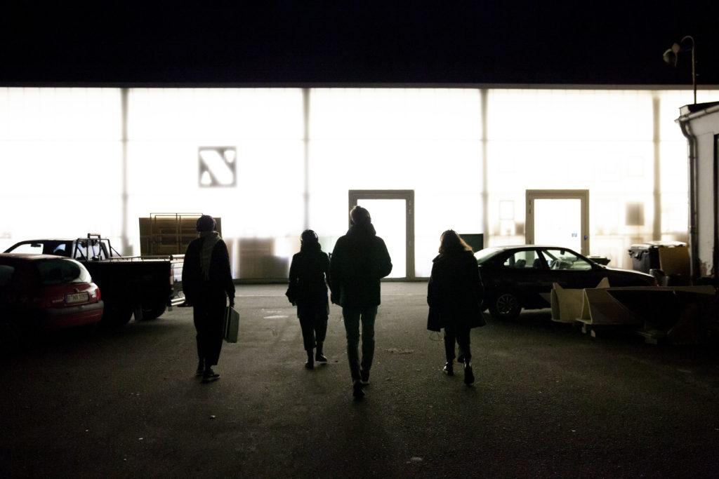 Four people walk across an unlit courtyard with parked cars toward brightly lit facades.