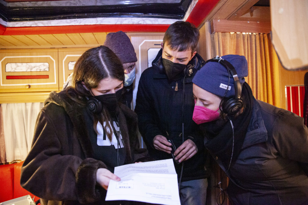 Four people wearing masks and headphones are standing in a motor home, reading from the same printed sheet of paper.