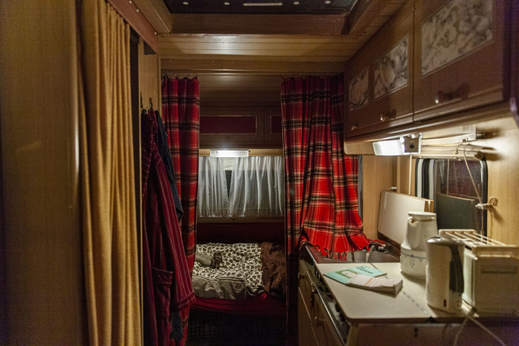 The interior of a motor home: a bed with brown and white sheets, checkered curtains, and a built-in kitchen with a toaster, an electric kettle, and a thermos.