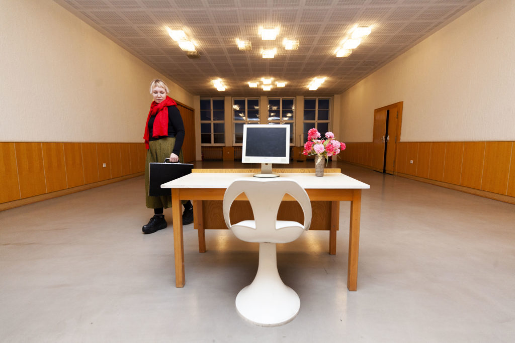 A large, elegant room with nothing in it except a table with a plastic chair, a screen, and flowers. A person with a briefcase approaches the table.