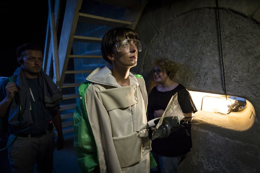 Eine Person mit Schutzbrille in einer Art Schutzanzug hält sich mit zwei anderen Person in Alltagskleidung in einem Kellergewölbe auf.