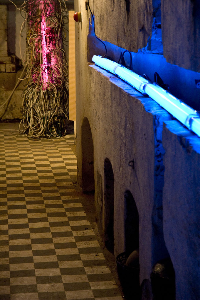 Ein Flur mit schwarzweiß gekacheltem Boden wird von in der Wand eingelassenen Leuchtstoffröhren beleuchtet. An seinem Ende steht ein pink leuchtendes Kabelgewirr.