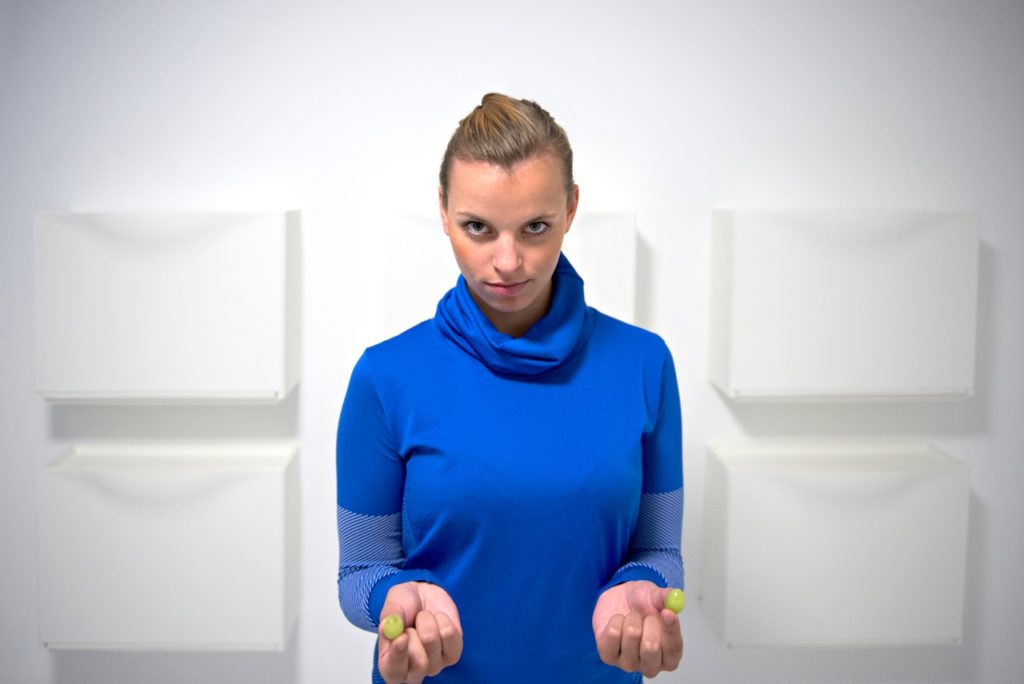 Eine Person in knallblauem Rollkragenpullover steht vor einer weißen Wand und hält in jeder Hand je eine Traube zwischen Daumen und Zeigefinger.