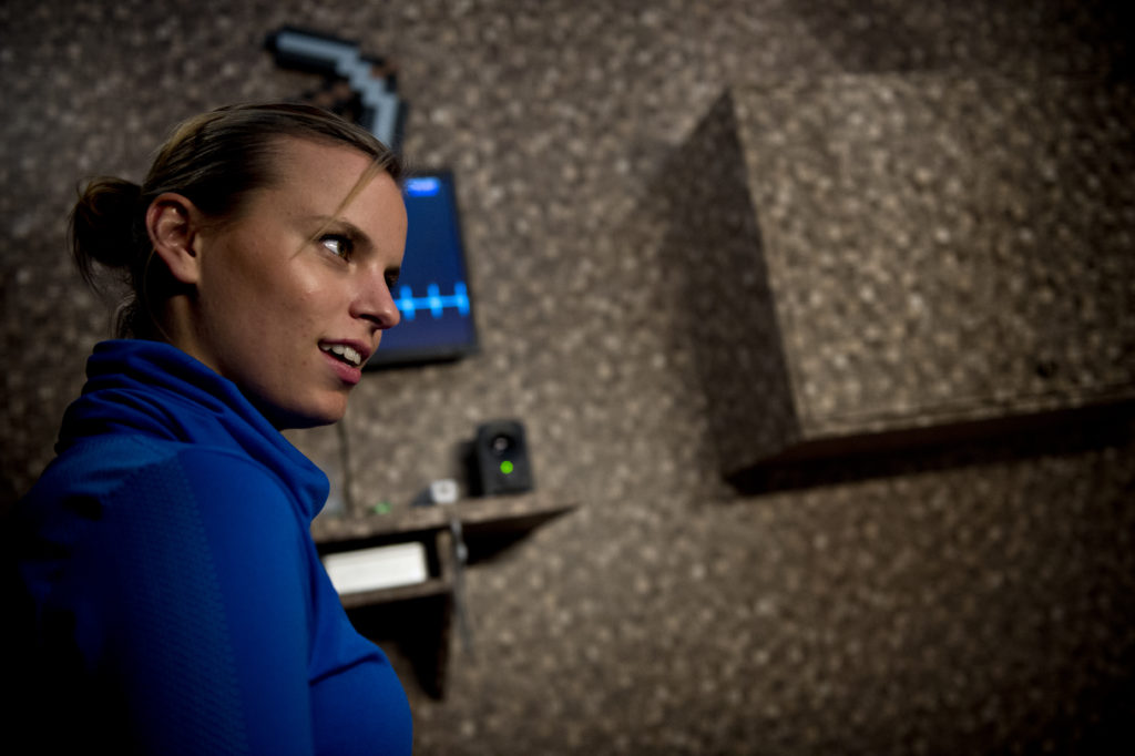 Eine Person mit strengem Dutt und knallblauem Oberteil steht vor einer Wand mit Bildschirm und Lautsprecher und schaut ernst an der Kamera vorbei.