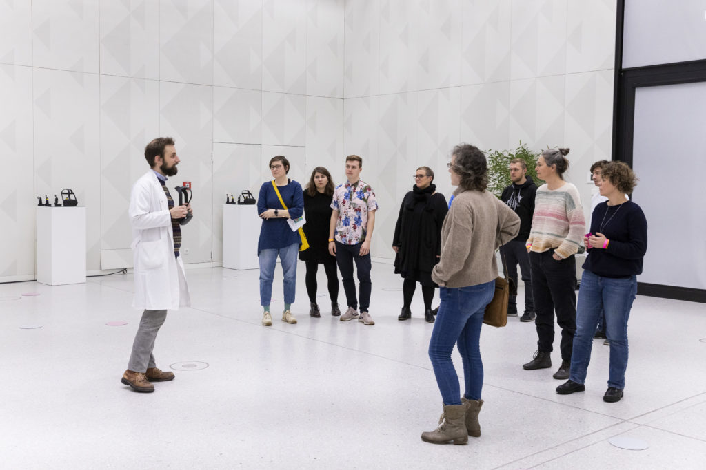 Eine Person in einem Laborkittel steht in einem komplett weißen Raum vor einer Gruppe Menschen und hält eine VR-Brille hoch.