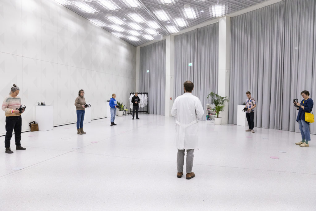 Eine Person in einem Laborkittel steht in einem komplett weißen Raum. Um ihn herum stehen Menschen und halten VR-Brillen in den Händen.