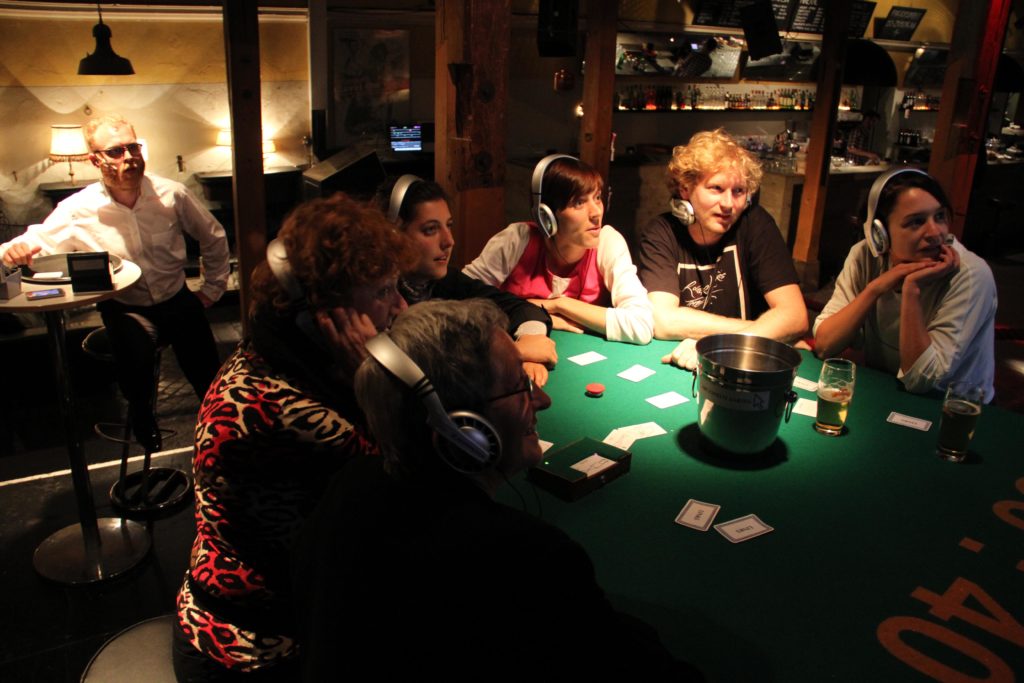 Menschen mit Kopfhörern sitzen an einem Casino-Tisch und schauen gebannt auf etwas. Im Hintergrund sitzt hinter einer Glasscheibe ein Mensch in Hemd.