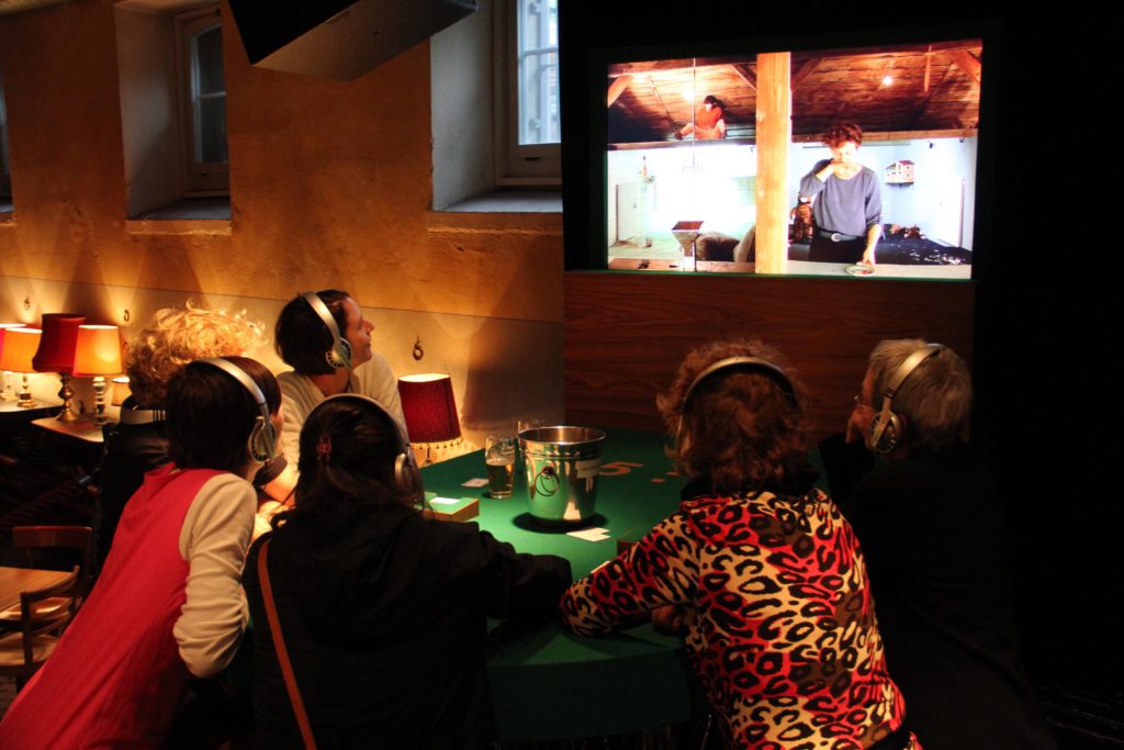 Personen an einem Casinotisch mit Kopfhörern schauen auf einem Bildschirm, auf dem zwei Personen in separaten Räumen zu sehen sind.