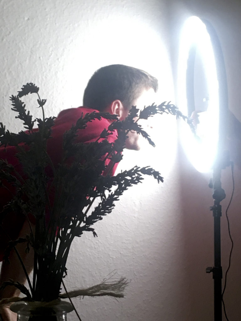 Eine Person starrt vorgebeugt in einen kreisförmigen Scheinwerfer auf einem Stativ. Sie wird von einer Zimmerpflanze im Vordergrund verdeckt.