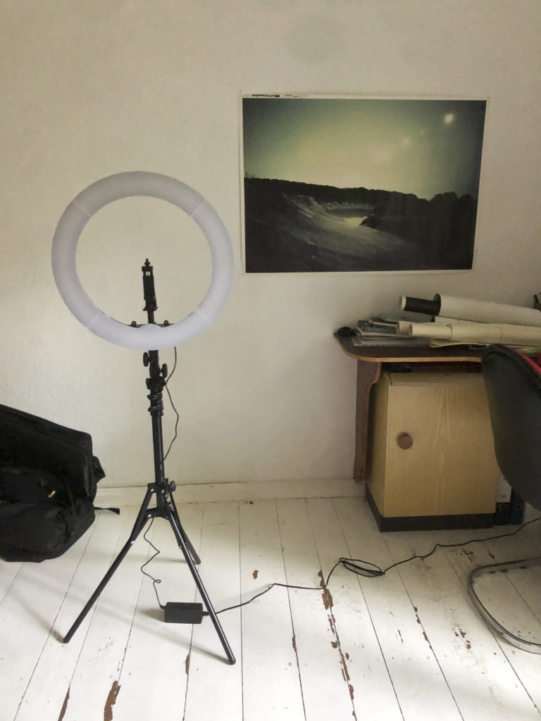 Ein kreisförmiges, ausgeschaltetes Licht auf einem Stativ steht auf einem Dielenbilden. Im Hintergrund ein Schreibtisch mit Papierrollen und ein Poster an der Wand mit einer Landschaftsaufnahme.