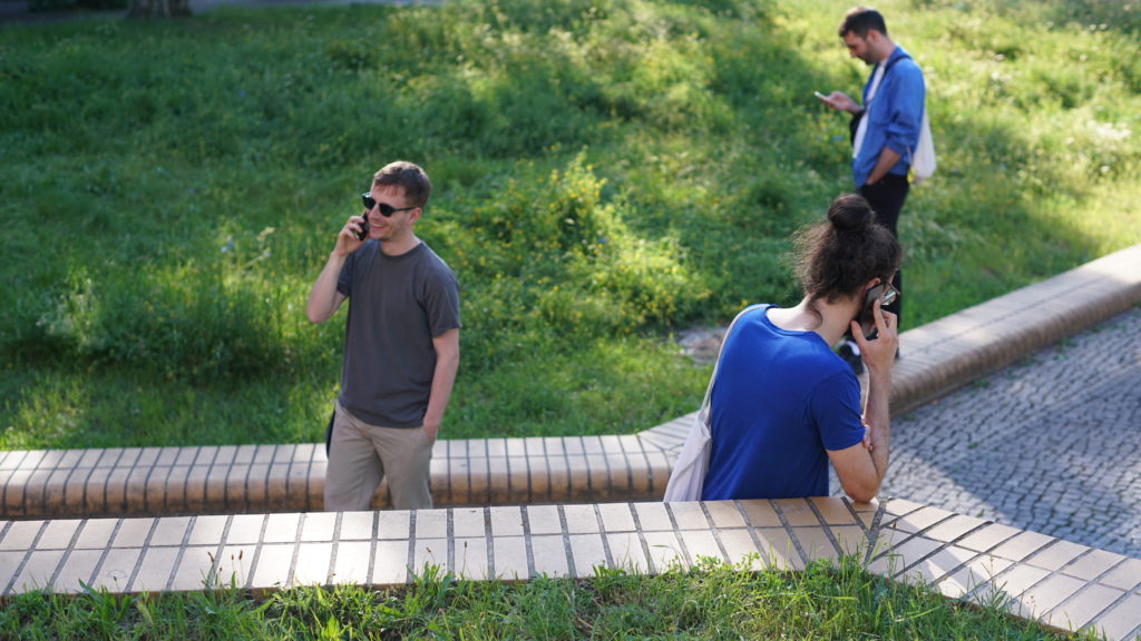 Drei Personen stehen in einer parkähnlichen Fläche herum und sind mit ihren Smartphones beschäftigt.