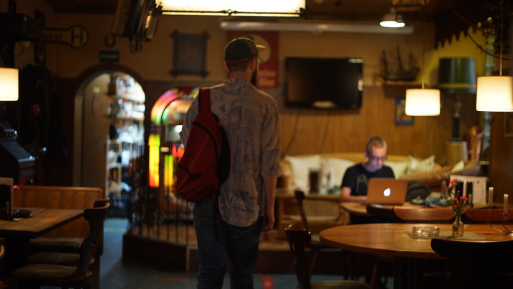 Eine Person mit Rucksack geht in einem Cafe auf eine Person an einem Tisch zu. Auf dem Tisch steht ein Macbook.