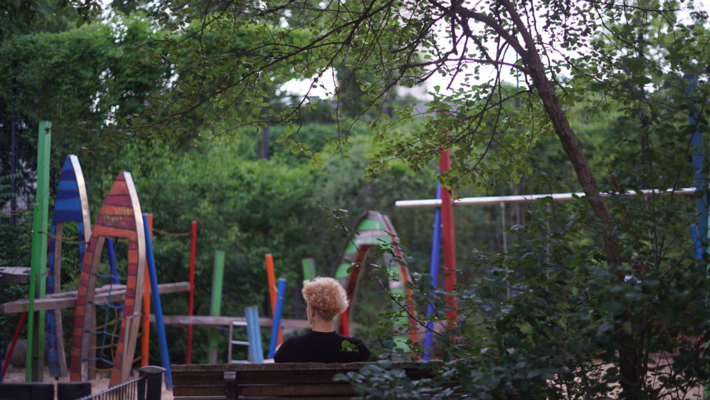 Eine Person sitzt auf einer Parkbank und schaut auf einen Spielplatz.