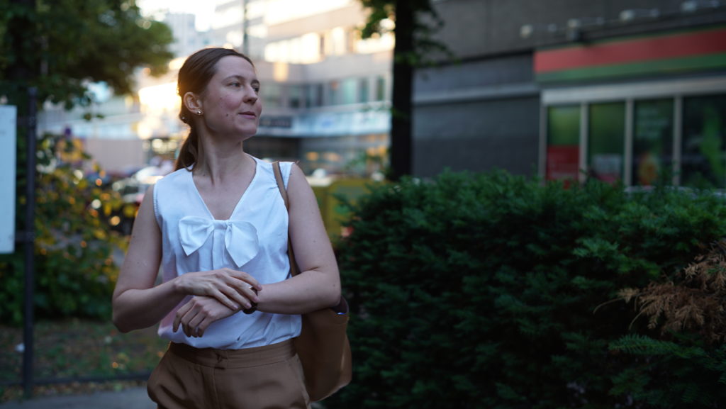 EIne Person in einer weißen ärmellosen Bluse mit einer beigen Handtasche steht vor urbanen Nachkriegsbauten und schaut rechts aus dem Foto heraus.