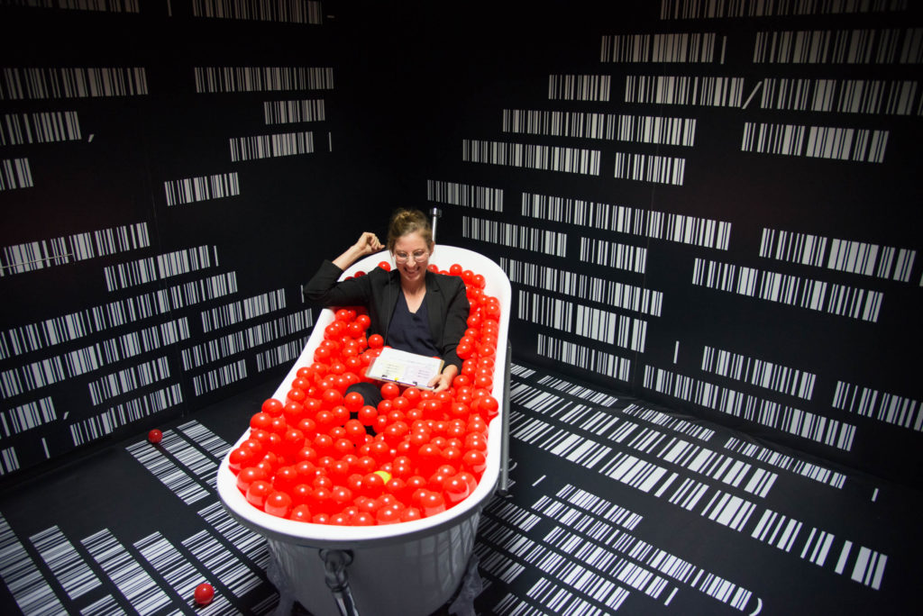 Eine Person sitzt mit einem Klemmbrett in einer Badewanne und lacht. Die Badewanne ist mit roten Plastikbällen gefüllt.