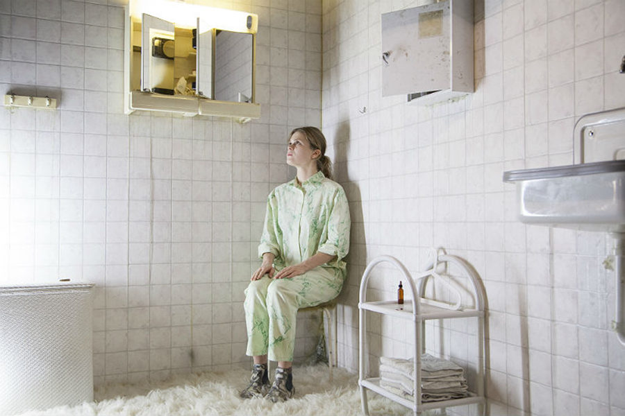 Eine Person in einem gemusterten Schlafanzug sitzt in einer Ecke in einem weiß eingerichteten Bad und schaut in die Ferne.
