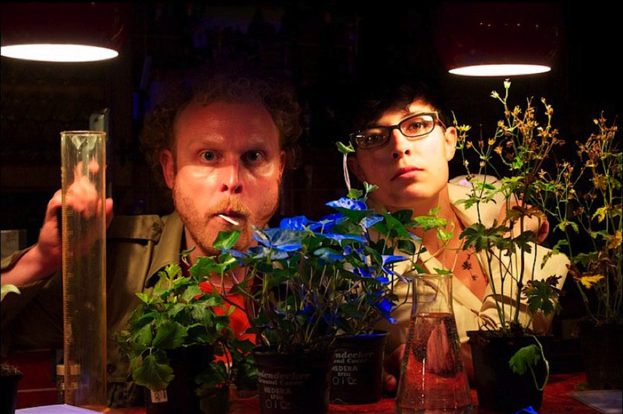Eine Person mit einem Lutscher im Mund und eine Person mit einer Sekretärinnen-Brille schauen in die Kamera. Vor ihnen auf einem Tisch stehen Pflanzen und Reagenzgläser.