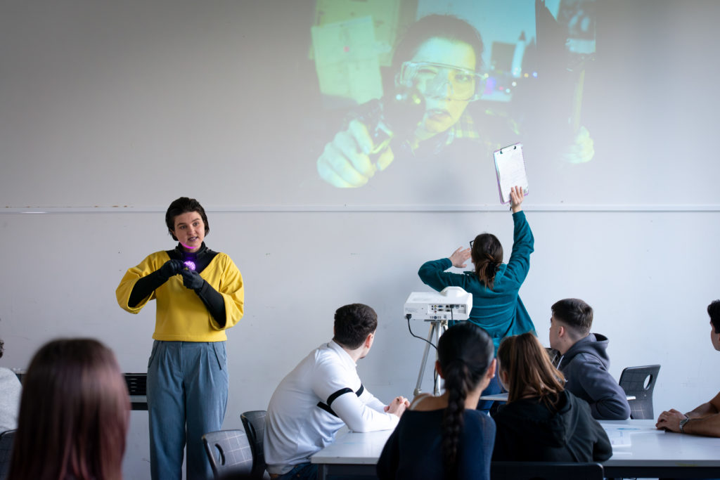 Eine als Android verkleidete Person und eine Wissenschaftlerin sowie die Projektion einer bastelnden Frau in einem Klassezimmer.